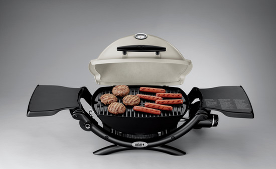 A portable charcoal grill with its lid opened, showing grilled burgers and hot dogs on the grate, set against a neutral background with a fireplace insert visible.
