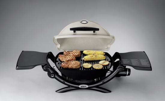 A weber electric grill opened to show cooked steak, corn, and slices of zucchini on the grate, with a gray background. The grill has a thermometer on the lid and side tables with a