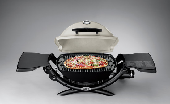 A portable electric grill open with a cooked pizza topped with various vegetables on the grilling surface, featuring a temperature gauge on the lid and fold-out side tables, and an insert for use as a fire