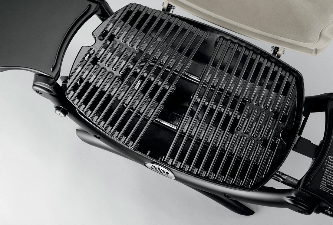 Close-up of a clean, black barbecue grill with metal grates and a Weber logo, viewed from above, with a portion of a beige grill cover on the left side and an insert for a fire