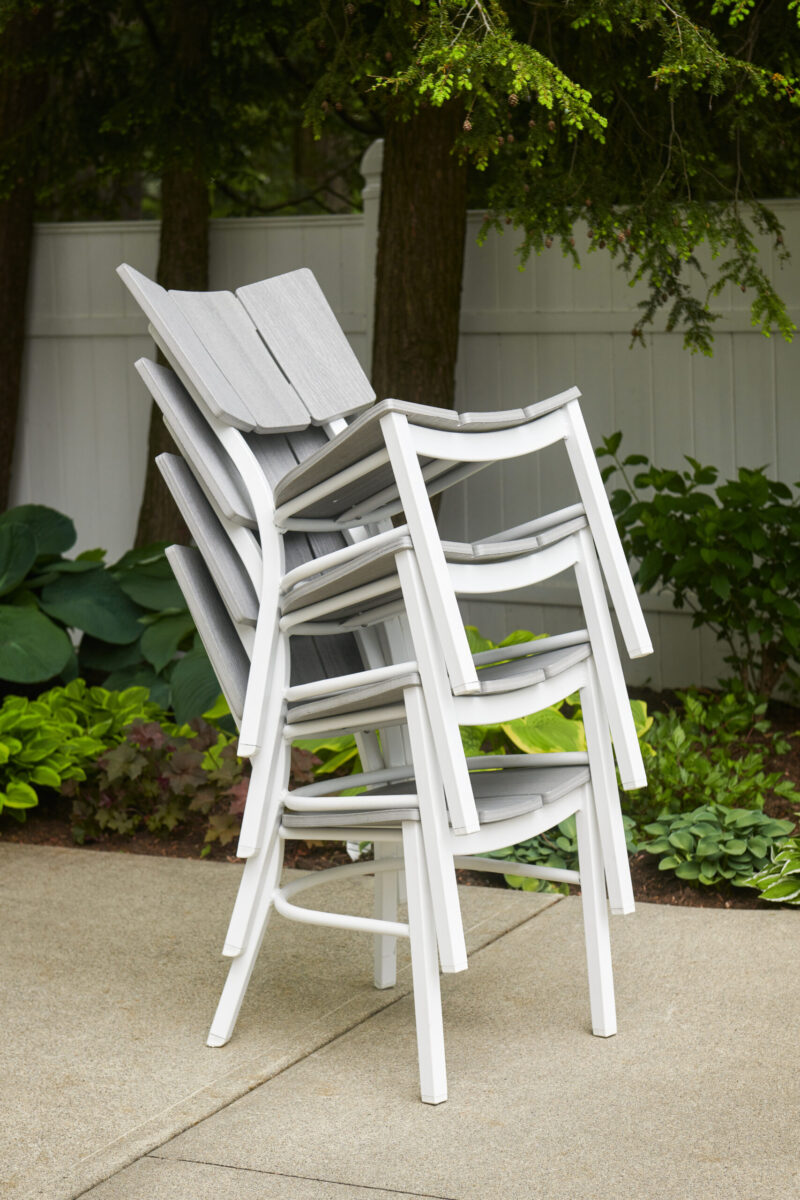 A stack of white plastic patio chairs placed on a concrete surface, with lush green plants and a white fence in the background.