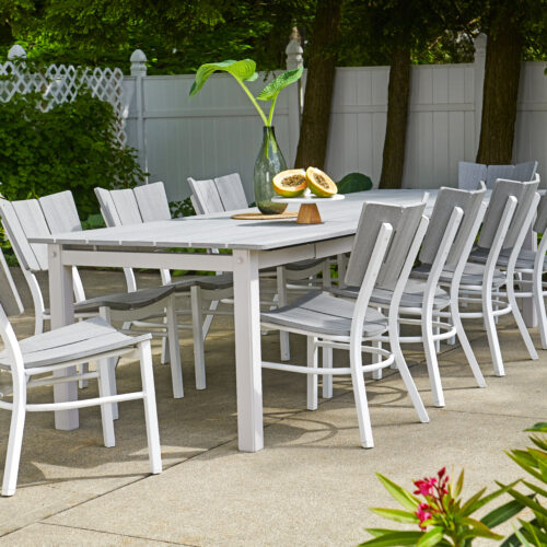 An outdoor dining area with a long white table and matching chairs set on a patio, surrounded by green plants, with a white fence in the background.