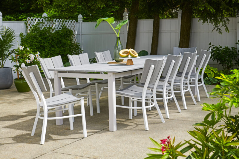 An outdoor dining area with a long white table and matching chairs set on a patio, surrounded by green plants, with a white fence in the background.