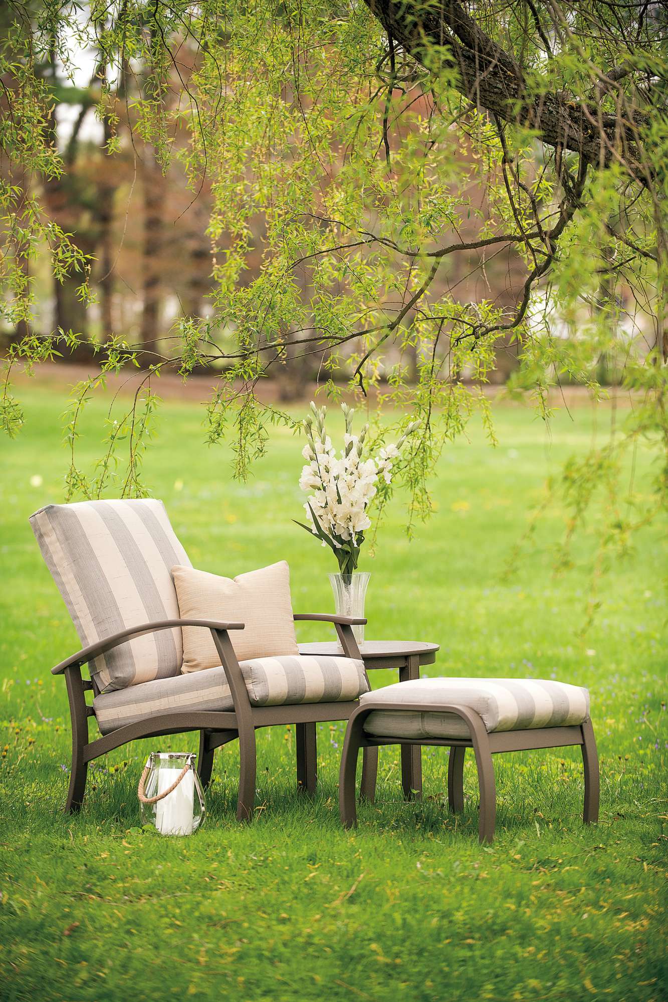 A cozy outdoor seating area with a cushioned chair and a matching footstool set on a lush green lawn, surrounded by springtime foliage and a fire pit with a vase of white flowers on a