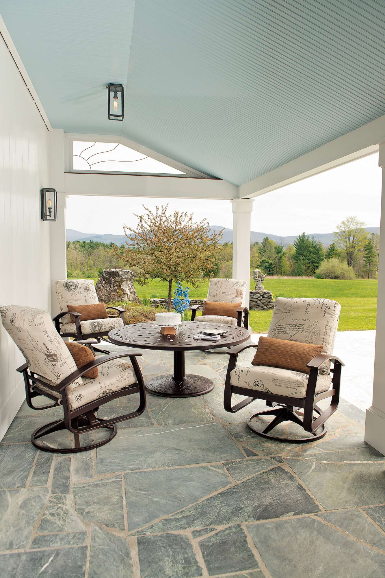 A covered outdoor patio features a set of four cushioned chairs around a round table with magazines and a fire pit, set on a stone floor, surrounded by lush greenery under a clear sky.