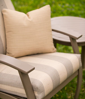 A close-up of an outdoor chair with striped cushions and a matching ottoman, set near a fire pit, against a blurred green grass background.