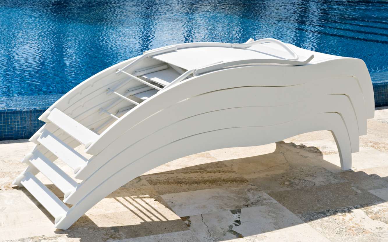 White modern curved lounge chair beside a swimming pool with clear blue water, set on a stone pool deck under bright sunlight, near a cozy fire pit.