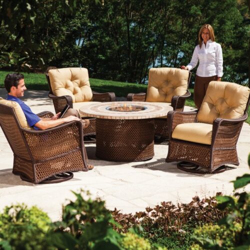 A man and a woman relax in a lush garden with outdoor wicker furniture, including plush chairs and a round table with a stone top, near a cozy fireplace, on a sunny day.