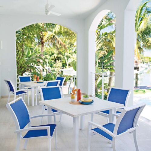 An airy, open patio dining area with white and blue chairs around a white table, set against a backdrop of lush tropical greenery and a clear sky, enhanced by the cozy addition of an outdoor fireplace