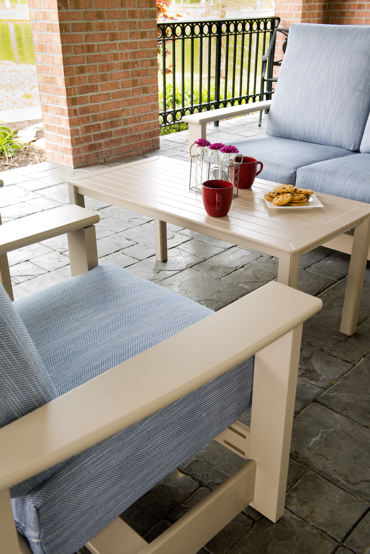 Outdoor patio area with a white table featuring a red pitcher and two cups, surrounded by cushioned white and blue chairs on a brick floor, under a covered porch with an insert fire pit.