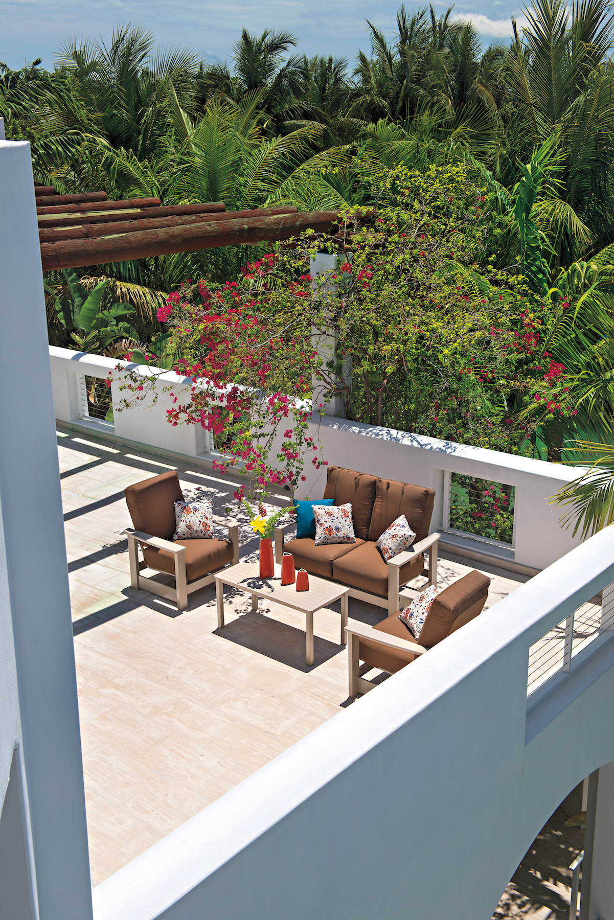 An outdoor terrace with modern furniture and vibrant floral arrangements overlooking a lush green landscape under a clear blue sky, featuring an inviting fire pit.