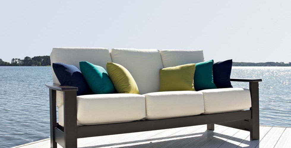 A modern white sofa with green and blue pillows on an outdoor deck overlooking a serene lake under a clear blue sky, next to a cozy insert fireplace.