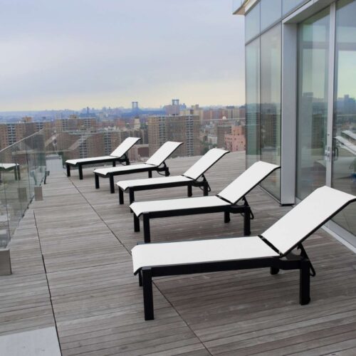 A row of white lounge chairs on a wooden deck with a glass barrier and an elegant fireplace, overlooking a cityscape with overcast skies.