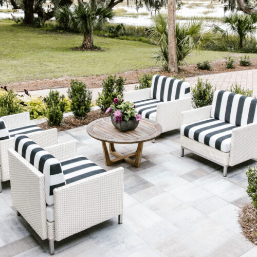 Elegant outdoor seating area with four wicker chairs outfitted in black and white striped cushions, centering around a wooden round table with an integrated fire pit, set against a backdrop of lush greenery