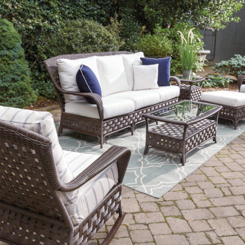 Outdoor patio furniture set with sofa, chairs, and coffee table on a rug, surrounded by lush greenery. The furniture is wicker with white cushions and blue pillows, featuring a central fire pit.