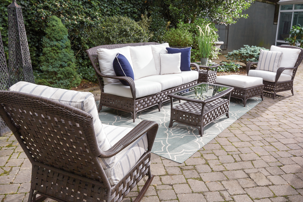 Outdoor patio furniture set with sofa, chairs, and coffee table on a rug, surrounded by lush greenery. The furniture is wicker with white cushions and blue pillows, featuring a central fire pit.