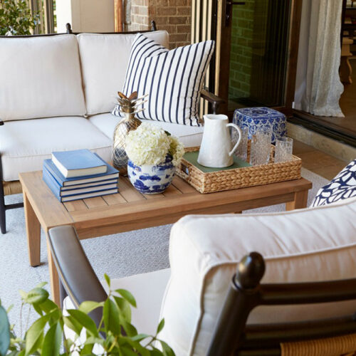 An inviting outdoor seating area with a plush white sofa adorned with blue and white striped cushions, a wooden Low Country coffee table displaying books and decorative items, and potted plants around.