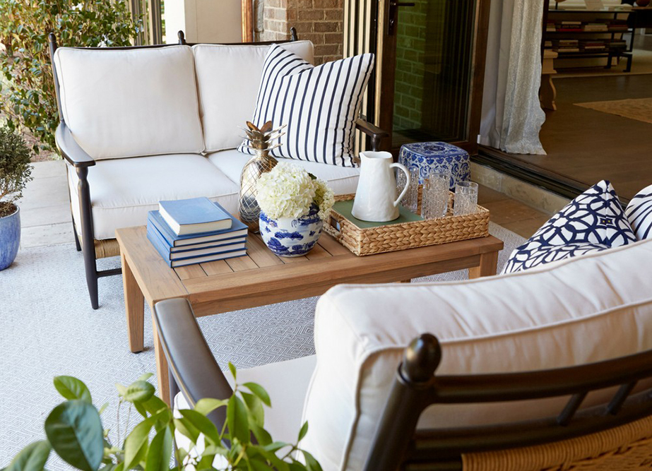 An inviting outdoor seating area with a plush white sofa adorned with blue and white striped cushions, a wooden Low Country coffee table displaying books and decorative items, and potted plants around.