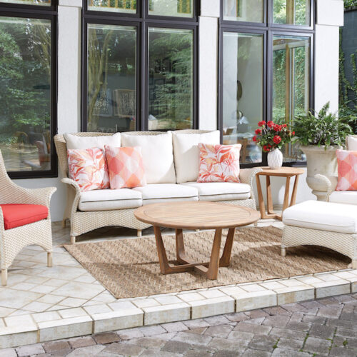 An elegant outdoor furniture setup on a patio, featuring a white sofa with coral pillows, two wicker chairs with one red cushion, a wooden coffee table, and a side table with an inserted vase of