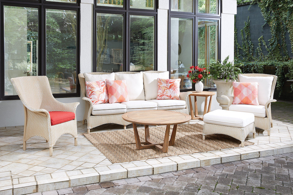 An elegant outdoor furniture setup on a patio, featuring a white sofa with coral pillows, two wicker chairs with one red cushion, a wooden coffee table, and a side table with an inserted vase of