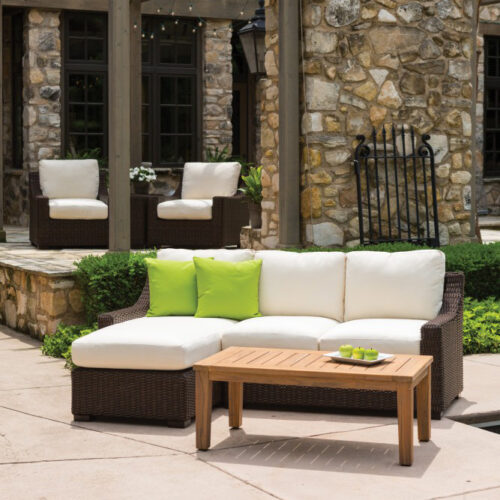 Outdoor patio with wicker furniture including a white couch with lime green pillows and two armchairs set against a stone building, complemented by a wood coffee table, greenery, and a fire pit