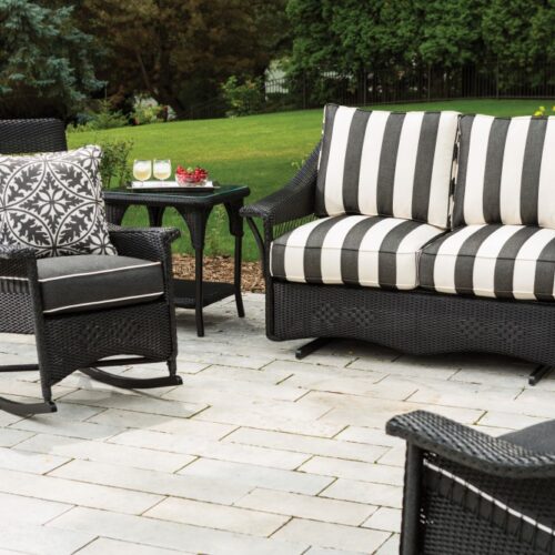 A cozy outdoor seating arrangement with a striped sofa and two chairs around a black side table, set on a patio overlooking a lush garden, complete with an inserted fire pit.