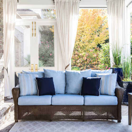 A bright sunroom with transparent curtains and a wicker sofa adorned with blue cushions, overlooking a garden with autumn foliage and a cozy fireplace insert.