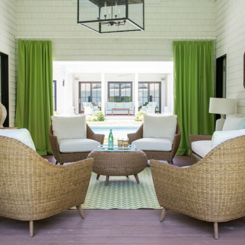 A bright, airy living room with lime green curtains, white furniture, and woven wicker chairs centered around a round wooden table. Large windows and plants add natural light and freshness to the space, enhanced