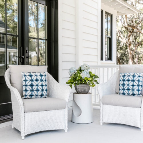 Two white wicker chairs with blue patterned cushions on a bright porch, featuring a center table with flowers and a nearby fire pit, near a house entrance surrounded by lush trees.