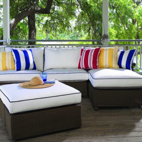 A spacious outdoor deck featuring a large l-shaped white cushioned sofa with blue and red striped and solid pillows, and an inserted fireplace nearby. A brown sun hat lies on the coffee table in the center
