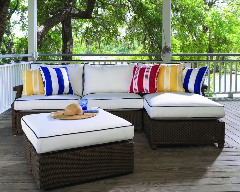 A spacious outdoor deck featuring a large l-shaped white cushioned sofa with blue and red striped and solid pillows, and an inserted fireplace nearby. A brown sun hat lies on the coffee table in the center