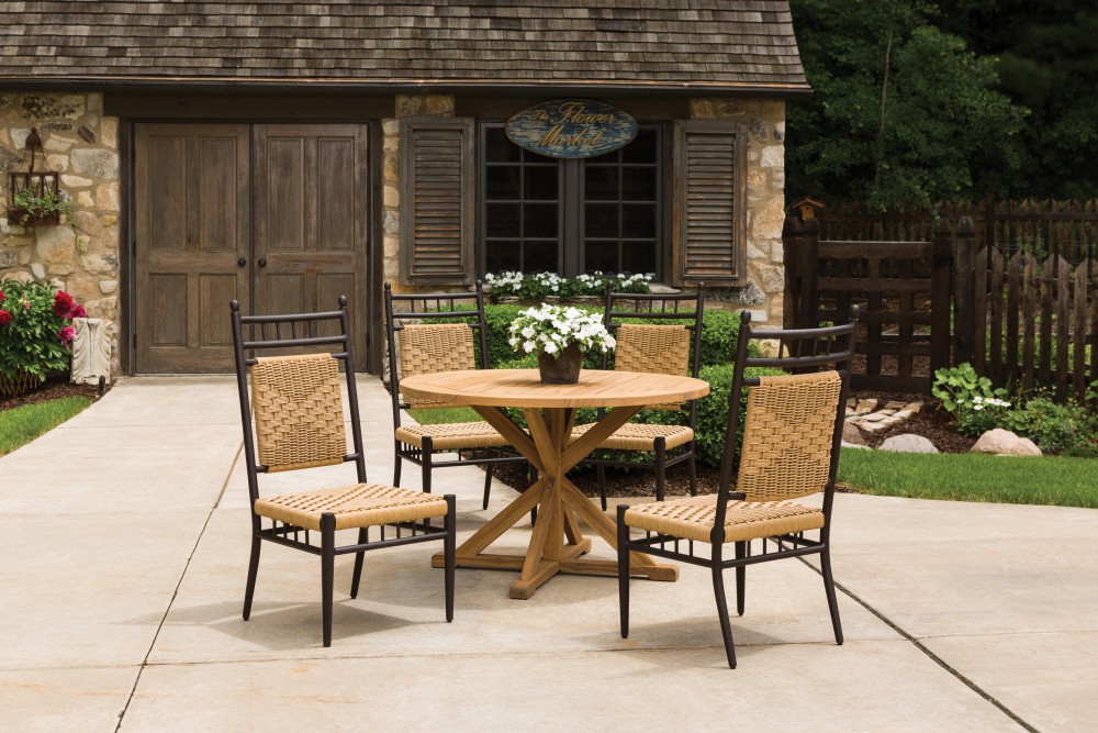 A cozy patio area featuring a round wooden table and four woven chairs on a stone pavement, with a charming rustic stone house and lush plants in the background. A stove insert adds warmth to this inviting outdoor