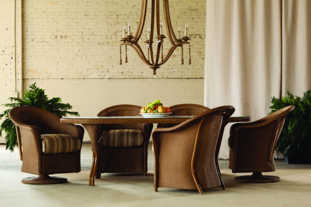 Elegant dining area featuring a round wicker table with four matching chairs, set against a cream curtain, brick wall, and a hanging chandelier, with green plants on either side and a fire pit