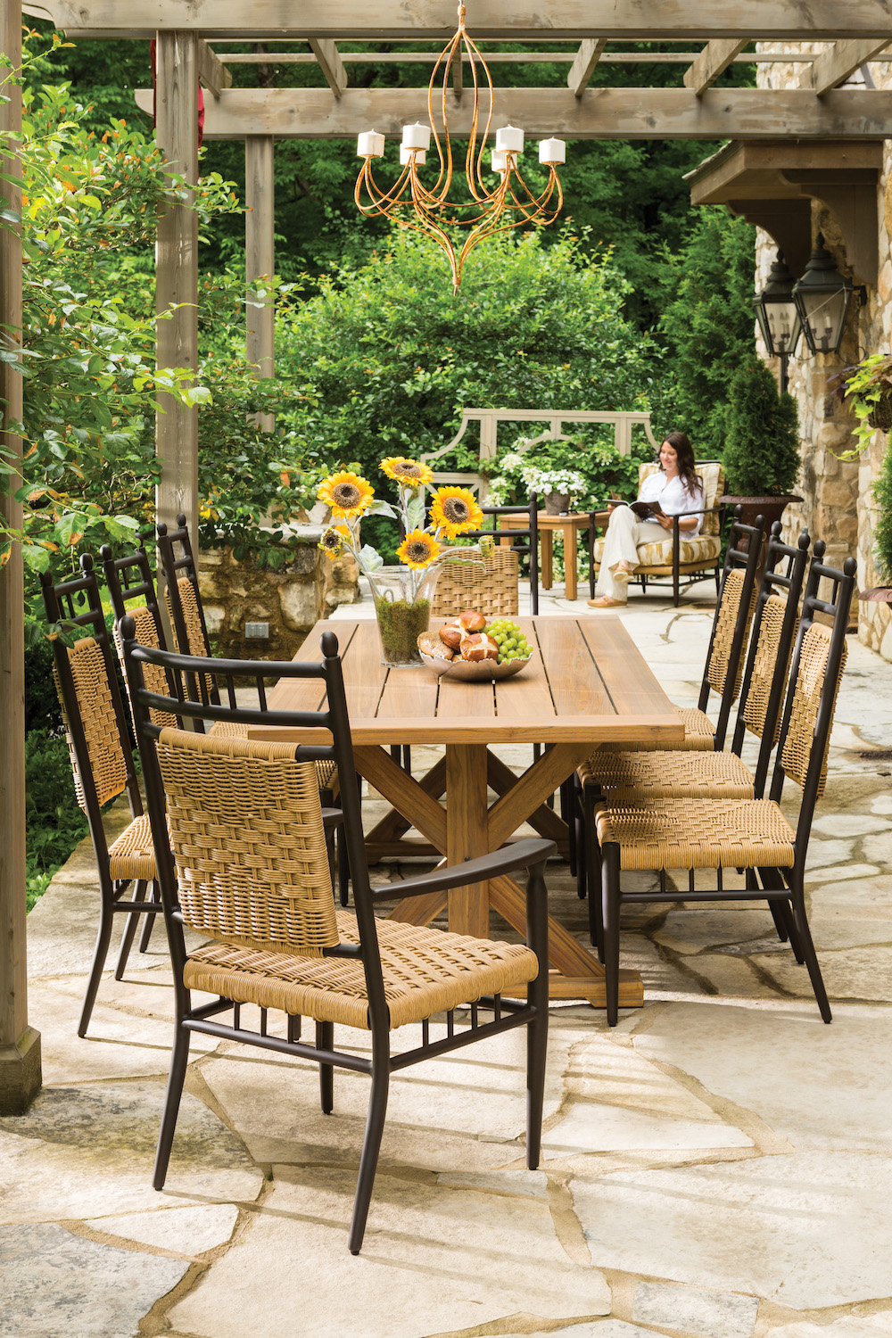 An elegant outdoor dining area featuring a wooden table with wicker chairs under a chandelier, surrounded by greenery. A fire pit is seen in the background with a woman sitting nearby.