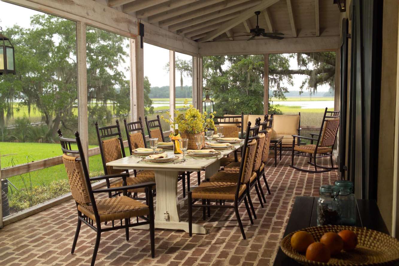 A cozy screened porch with a dining setup overlooking a serene landscape, featuring wicker chairs, a wooden table set with dishes, and a fire pit amidst the lush greenery in the background.