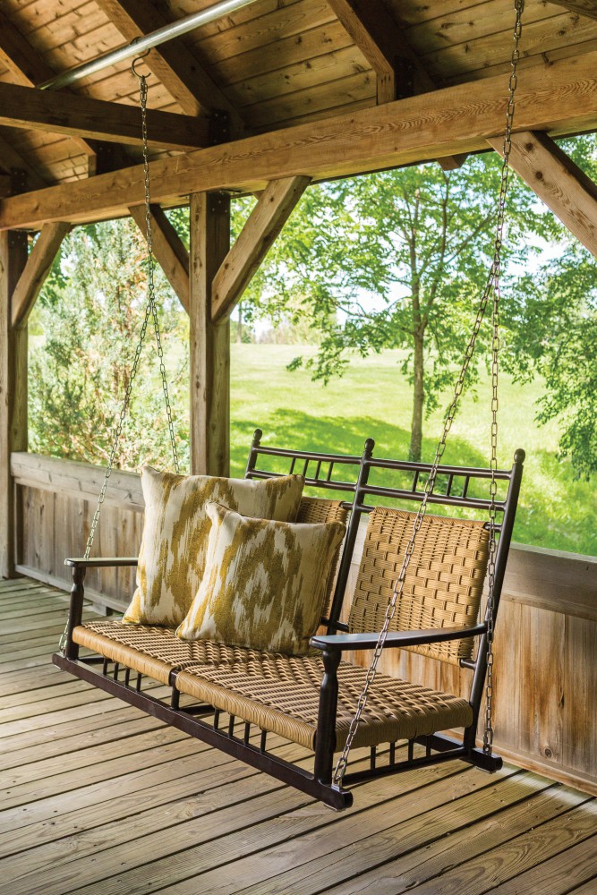 Wooden porch swing with plush cushions, hanging from a beam with chains, set against lush greenery and a cozy fireplace in the background on a sunny day.