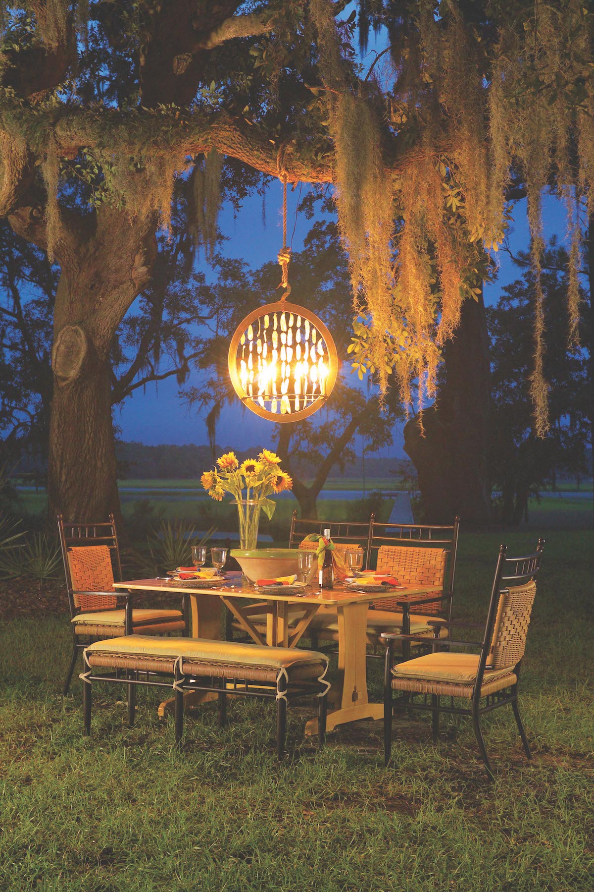 An outdoor dining area under a large tree with hanging Spanish moss, lit by a round light fixture at dusk. The table is set for a meal and surrounded by chairs, featuring a fire pit in the