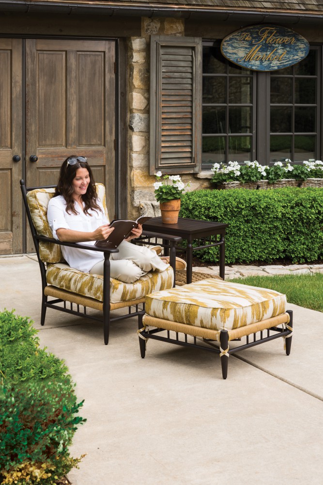 A woman smiling, reading a magazine while reclining on a cushioned lounge chair outside a stone house with plants, a fireplace, and a "home sweet home" sign.