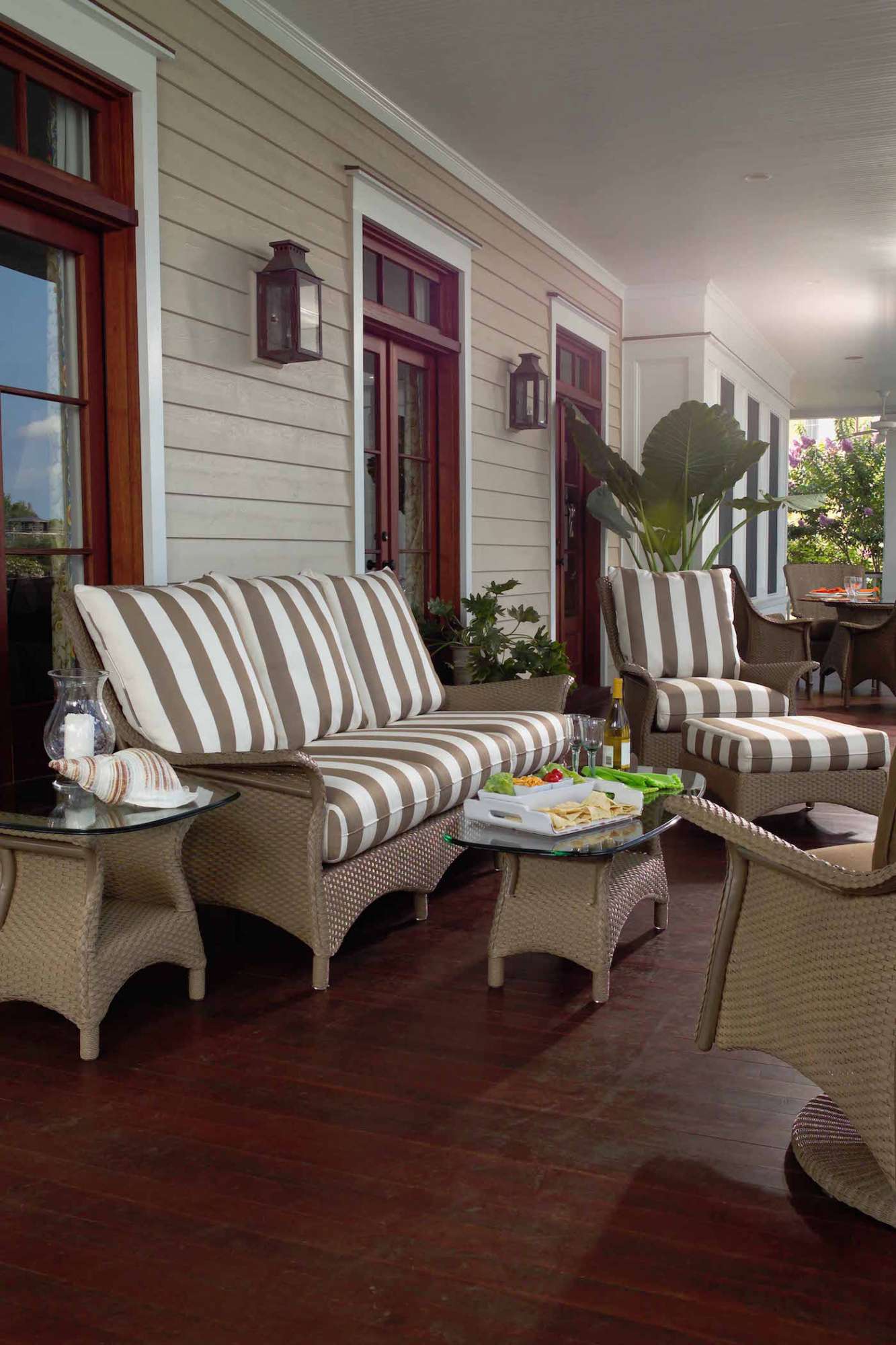 A cozy porch featuring striped cushioned chairs and a sofa arranged around a central coffee table with books and a plant, flanked by large potted plants near the entryway, enhanced with an inserted fire