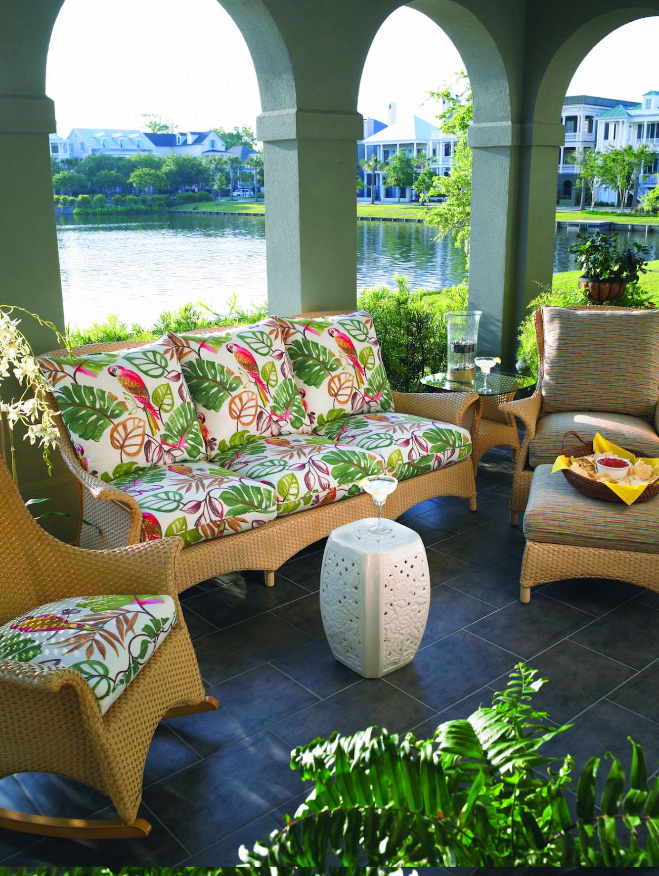 An inviting patio with wicker furniture, including a patterned sofa and chair, set under archways overlooking a serene river. A cozy fire pit enhances the tranquil, relaxing outdoor setting with lush plants.
