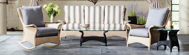 An elegant patio furniture set featuring two armchairs and a sofa with striped cushions, centered around a glass-top coffee table, next to a fire pit in a warmly lit, open-air setting.