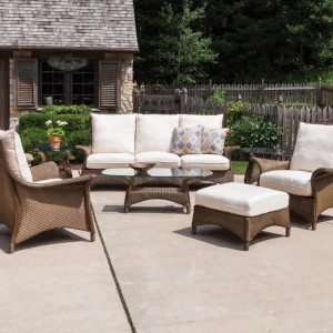 An outdoor patio furnished with wicker furniture, including a sofa, chairs, and a coffee table, set on a flagstone floor with a cozy cottage background and a wooden fence featuring an insert fire pit
