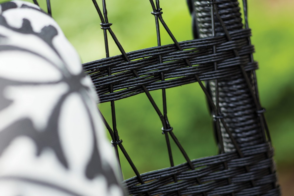 Close-up view of a black wicker chair with a detailed pattern, partially obscured by a blurred foreground of patterned fabric near a fireplace.