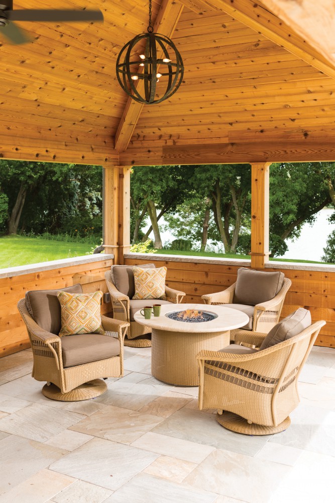 Cozy outdoor seating area under a wooden gazebo with a fireplace table surrounded by wicker chairs with cushions, overlooking a lush green landscape.