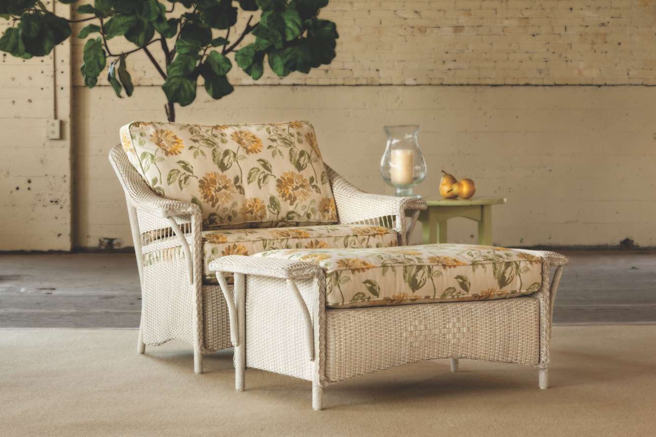 A cozy living room setup featuring a wicker loveseat and chair with floral cushions, against a white brick wall. A fireplace insert is visible beside the arrangement. A side table holds a pitcher and glass