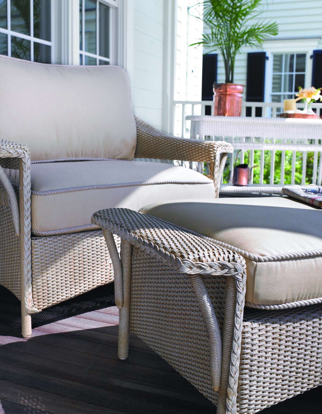 Two woven wicker chairs with plush beige cushions on a wooden deck, overlooking a railing with a view of a white house with black shutters and a fire pit.