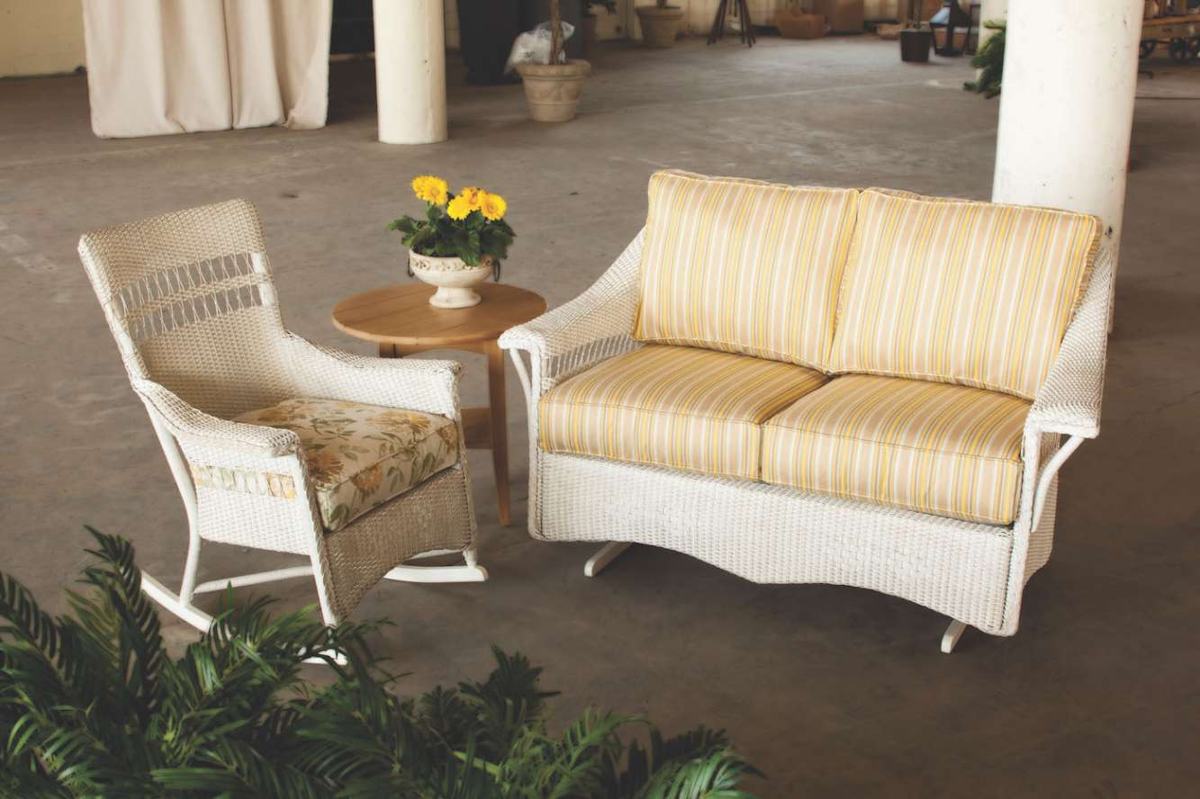 Two wicker chairs with cushions and a small wooden table, set in a spacious room with potted plants near a fireplace, creating a cozy seating area.
