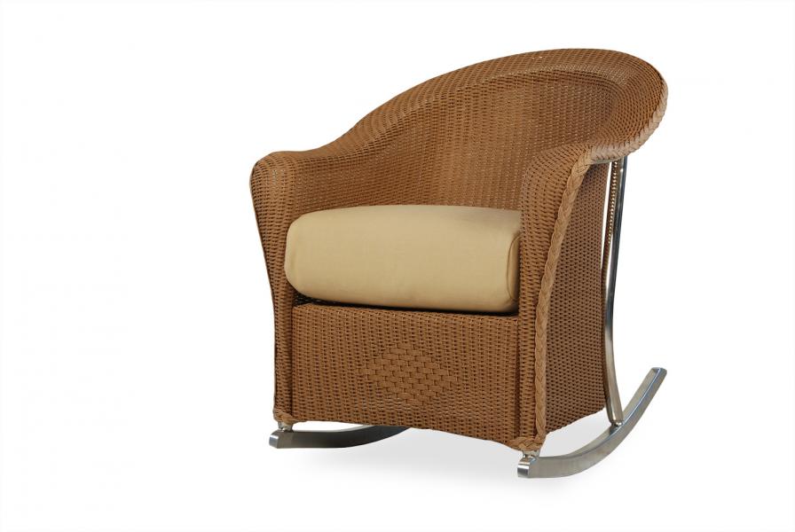 A modern rocking chair made of brown wicker with a light beige cushion, viewed from the side, against a white background near a fireplace.
