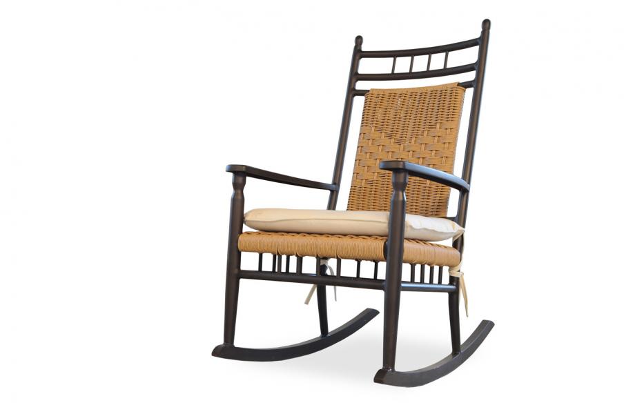 A traditional rocking chair with a dark wood frame and woven rattan backrest, featuring a beige cushion on the seat, and placed near a fireplace, against a white background.