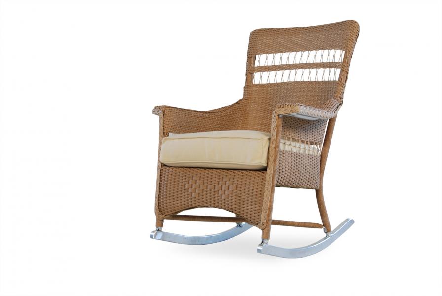 A modern wicker rocking chair with a beige cushion on a white background. The chair features a high back, armrests, and sleek metal runners, perfectly positioned near a fireplace.